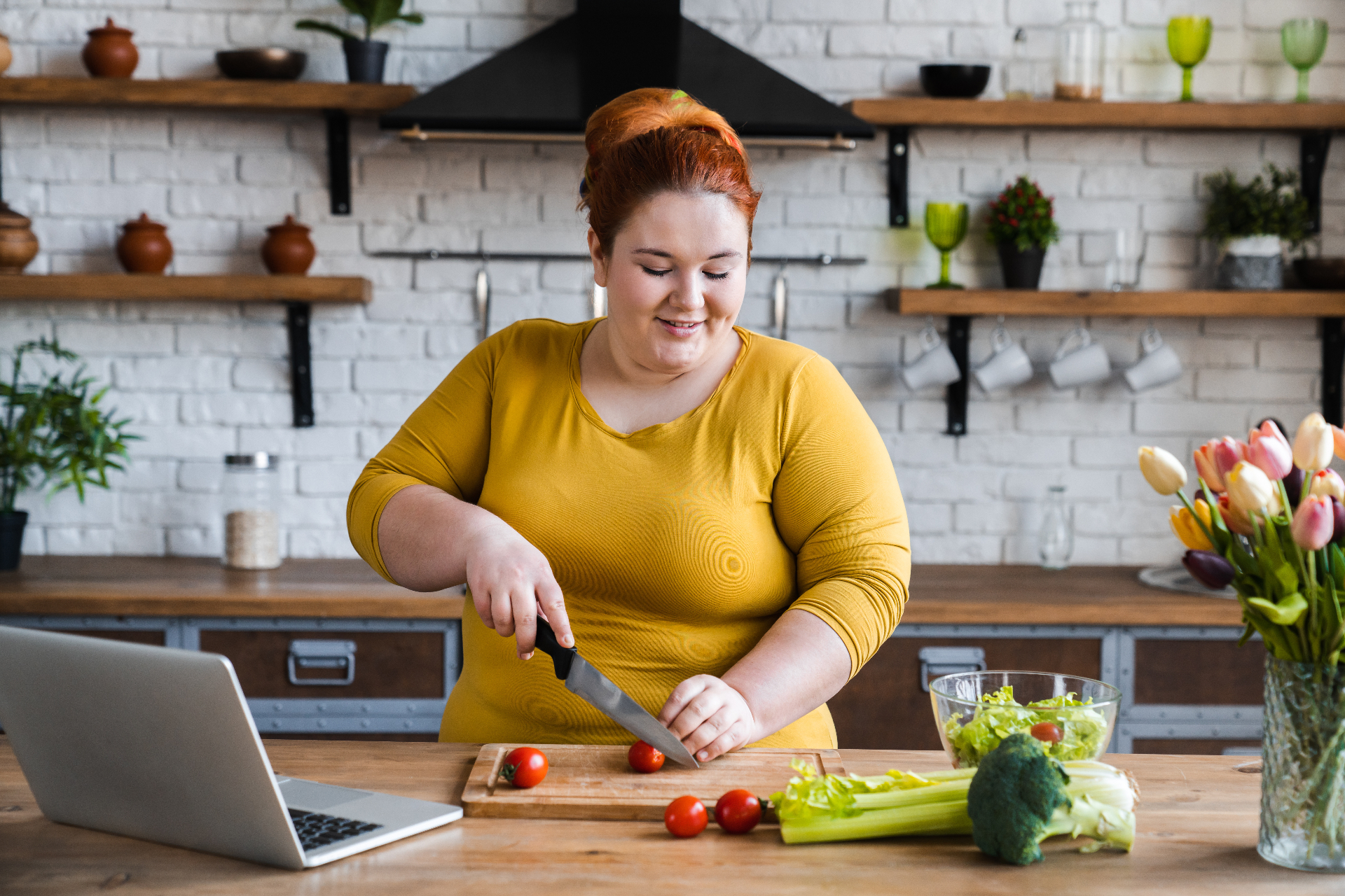 Smiling woman chopping fresh tomatoes and vegetables in a bright kitchen with a laptop nearby